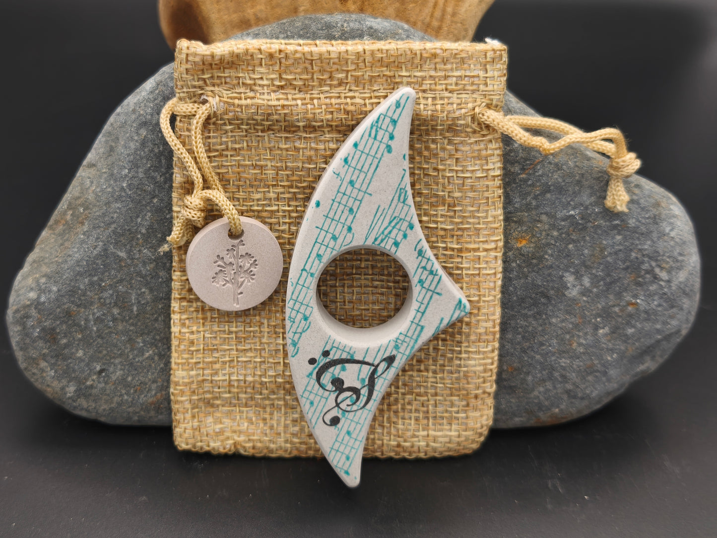 Bague de lecture artisanale gris clair décorée d’une mélodie turquoise et d’une clé de sol noire, présentée sur un pochon en toile de jute – création Omiralys.