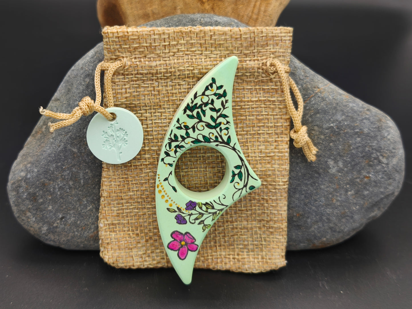 Bague de lecture artisanale vert amande décorée de lianes florales noires et d’une fleur rose, présentée sur un pochon en toile de jute – création Omiralys.