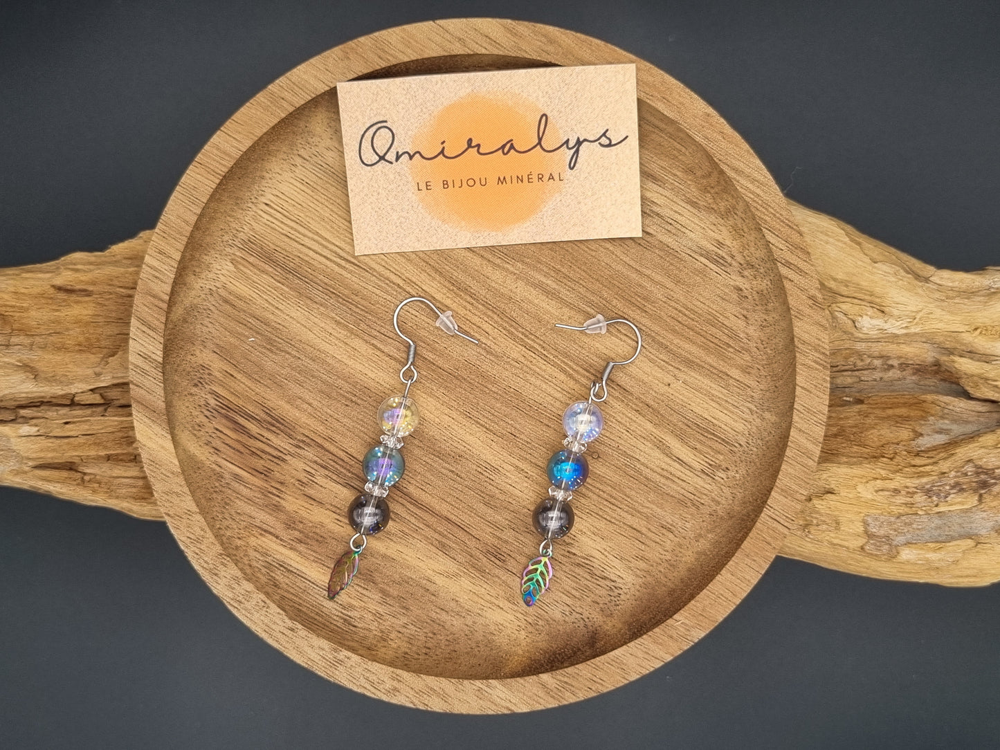 Boucles d'oreilles plumes Omiralys en acier inoxydable avec perles Aqua Aura irisées présentées sur un plateau en bois artisanal.