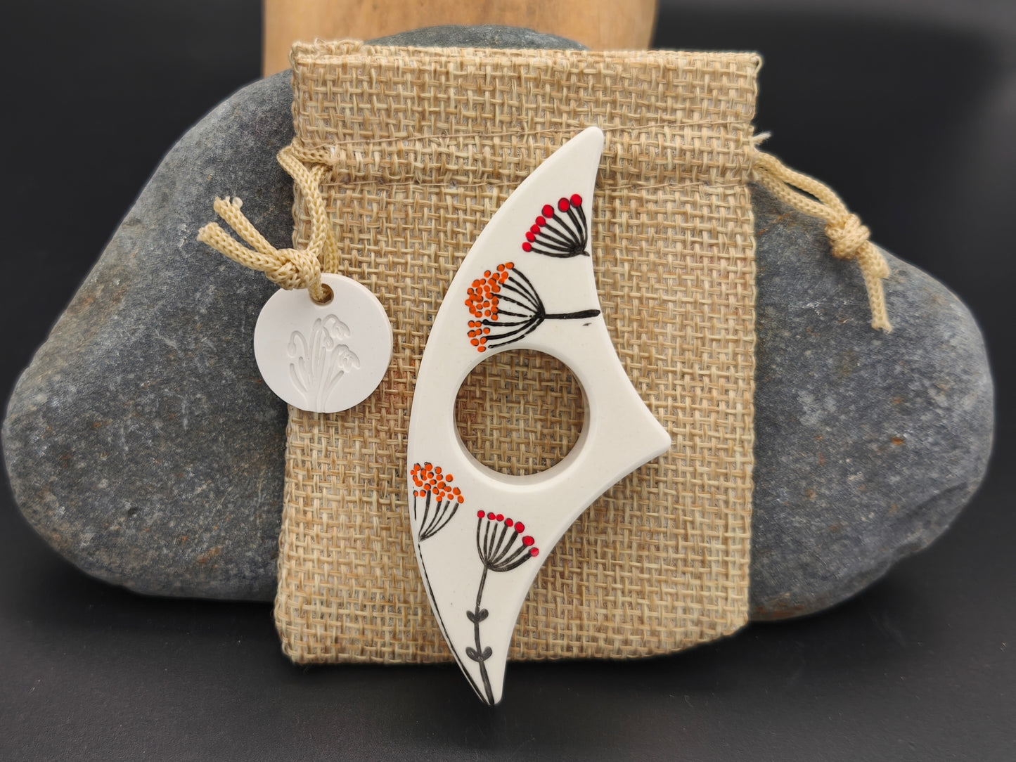 Bague de lecture artisanale Omiralys blanche avec fleurs stylisées orange, rouge et noir sur son pochon en toile de jute.