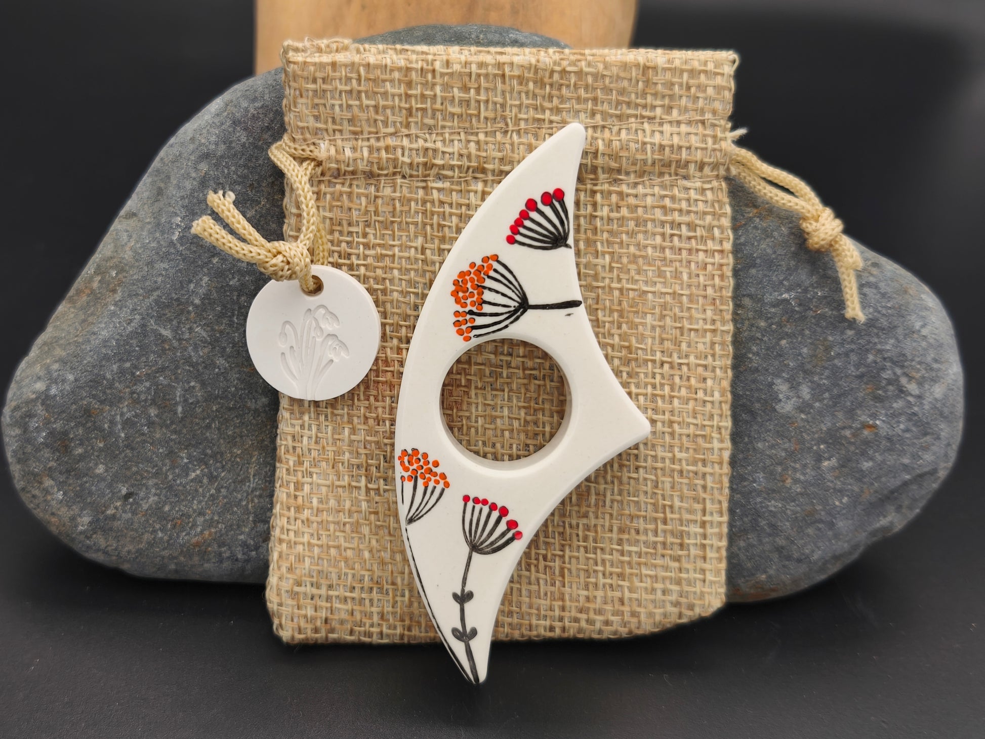 Bague de lecture artisanale Omiralys blanche avec fleurs stylisées orange, rouge et noir sur son pochon en toile de jute.
