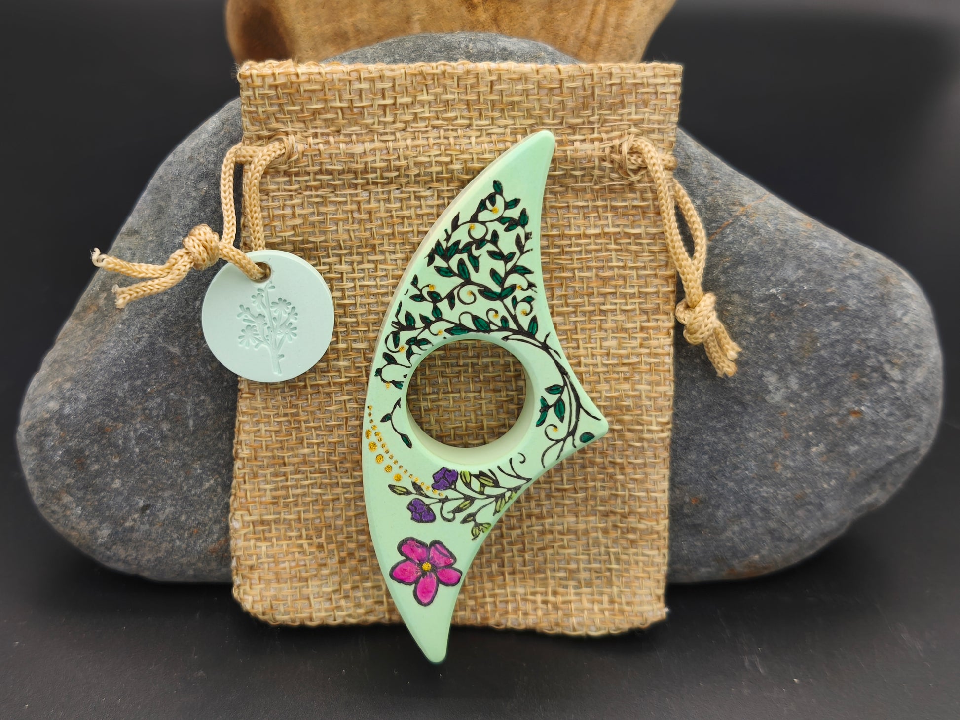 Bague de lecture artisanale Omiralys vert amande avec décor lianes noires et fleur rose sur son pochon en toile de jute.