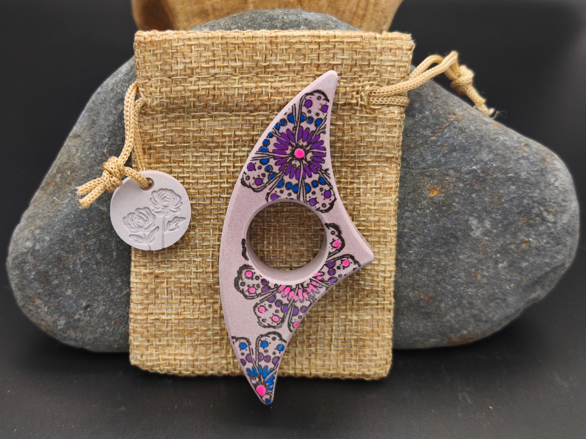 Bague de lecture artisanale mauve rosé décorée d’un mandala floral et de pétales violets, présentée sur un pochon en toile de jute – création Omiralys.