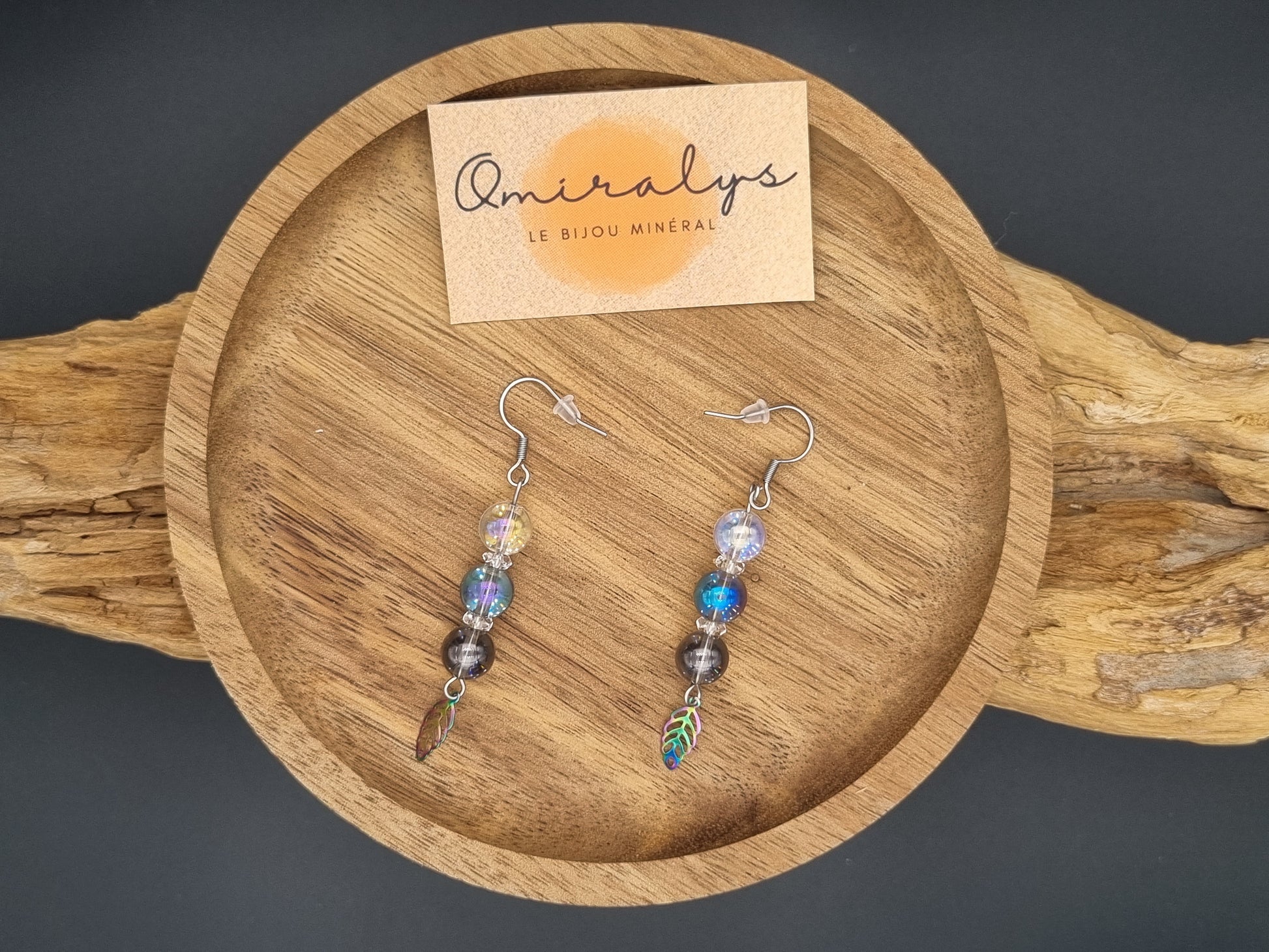 Boucles d'oreilles plumes Omiralys en acier inoxydable avec perles Aqua Aura irisées présentées sur un plateau en bois artisanal.